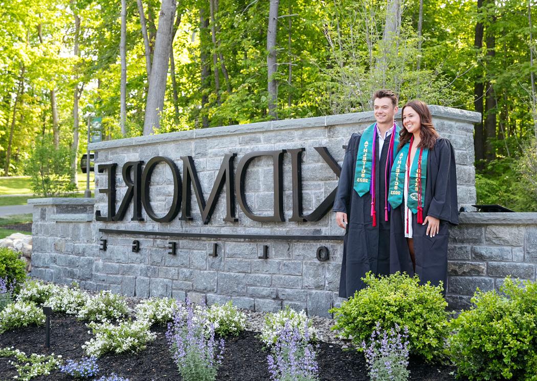 two college graduates post in front of a 校园 entrance sign that says Skidmore College